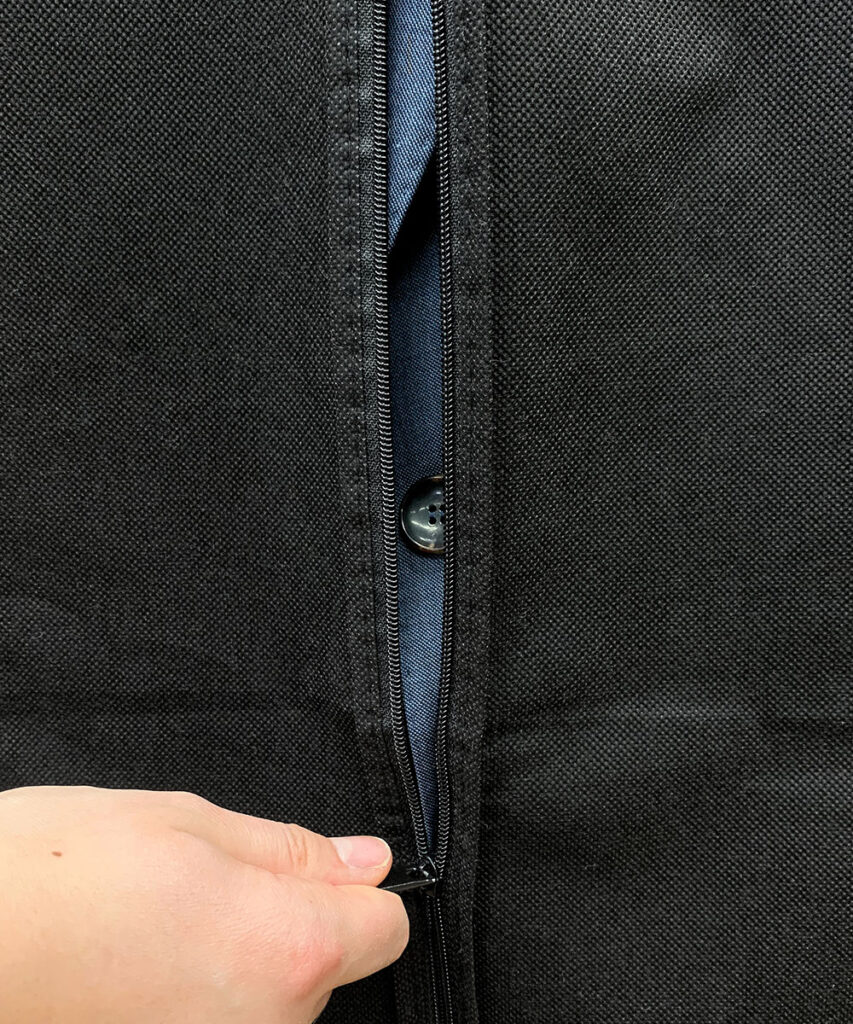 Deailaufnahme eines schwarzen Kleidersackes, dessen Reißverschluss von einer Hand zugezogen wird, im Kleidersack erahnt man einen blauen Woll-Anzug
