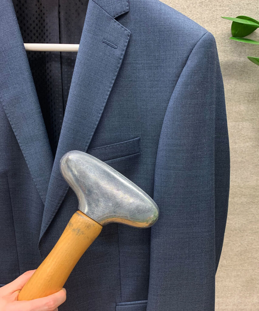 Blauer Woll-Anzug auf Bügel vor beigem Hintergund, welcher mit einem metallenen Steamer gesteamt wird; rechts im Hintergund eine Zimmerpflanze