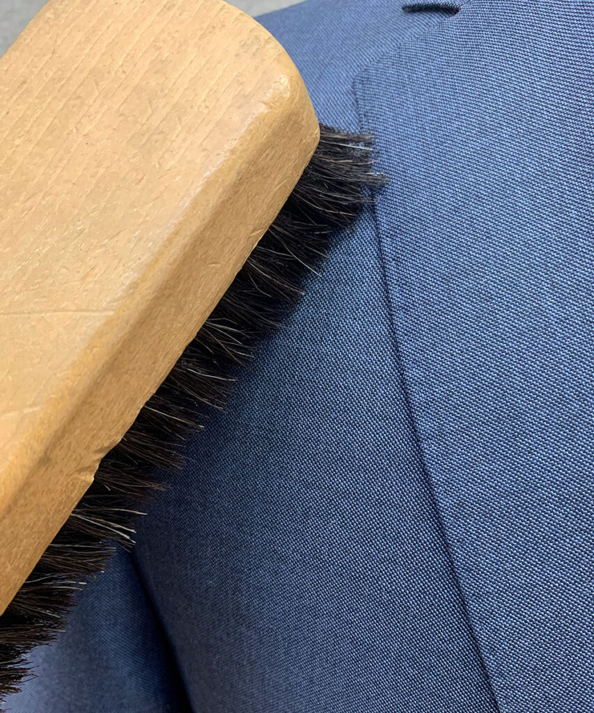 Detailaufnahme eines blaen Woll-Anzuges, welcher mit einer Naturhaarbürste aus Holz mit schwarzen Borsten ausgebürstet wird