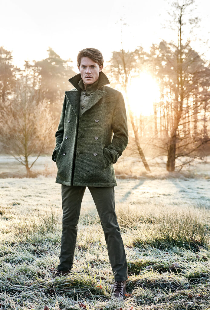 young man with brown hair wearing green trousers and a green, structured wool coat, looking directly into the camera. In the background a heath landscape, one sees bare trees through which the sun shines.