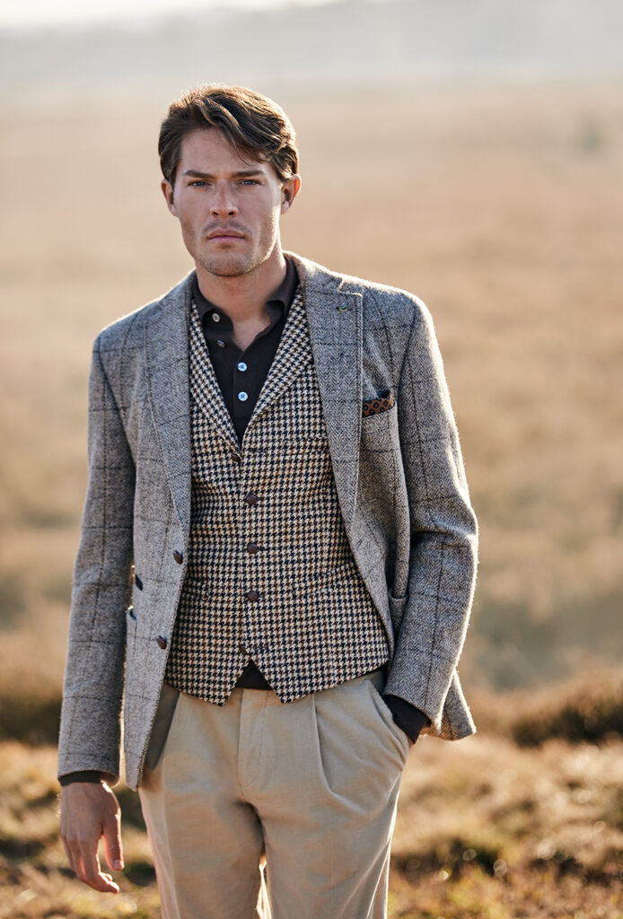 man with brown hair in brown polo, beige trousers, checked waistcoat and checked jacket looking at the camera, a heath landscape in the background out of focus.