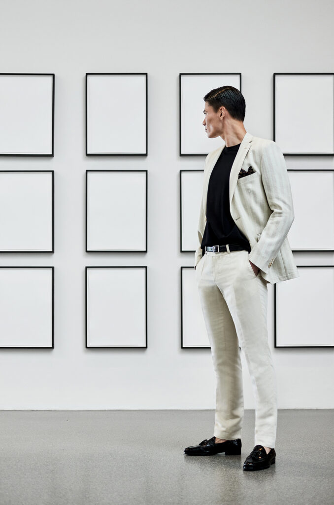 Young man with dark hair in black shirt, white suit trousers with black belt and white jacket. He is wearing black suit shoes and has both hands in his trouser pockets. In the background you see a white wall with 12 white pictures in black frames.