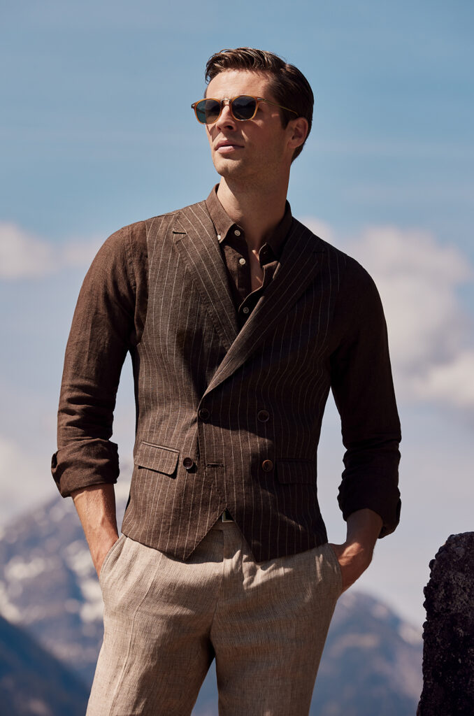 Man with brown hair and sunglasses in brown shirt, beige trousers and brown striped waistcoat. He has both hands in his trouser pockets. In the background you can see the top of a mountain and a blue sky with clouds.