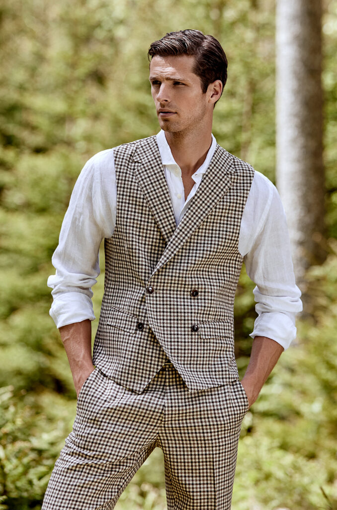 Man with brown hair in a white shirt, black and white checked waistcoat and black and white checked trousers. He has both hands in his trouser pockets and is looking towards the screen. in the background you can see a forest landscape. 