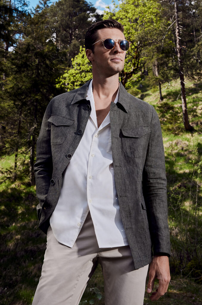 Man with brown hair and sunglasses in white shirt, brown shirt jacket and beige trousers. In the background you can see a forest landscape. 