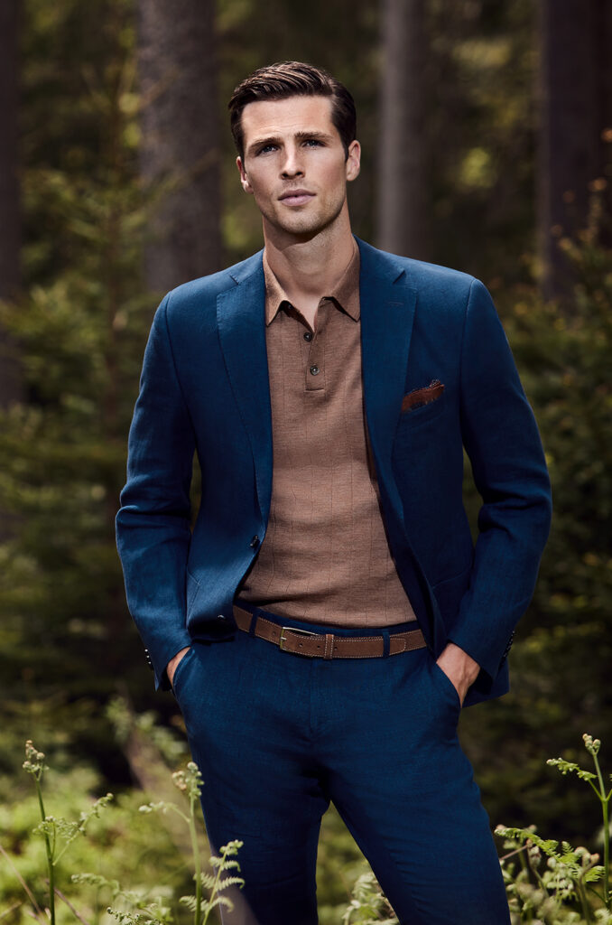 Young man with brown hair in brown shirt and blue GOTS certified CONCEPT GREEN suit. He looks directly into the camera and has both hands in his trouser pockets. In the background you can see a forest landscape. 