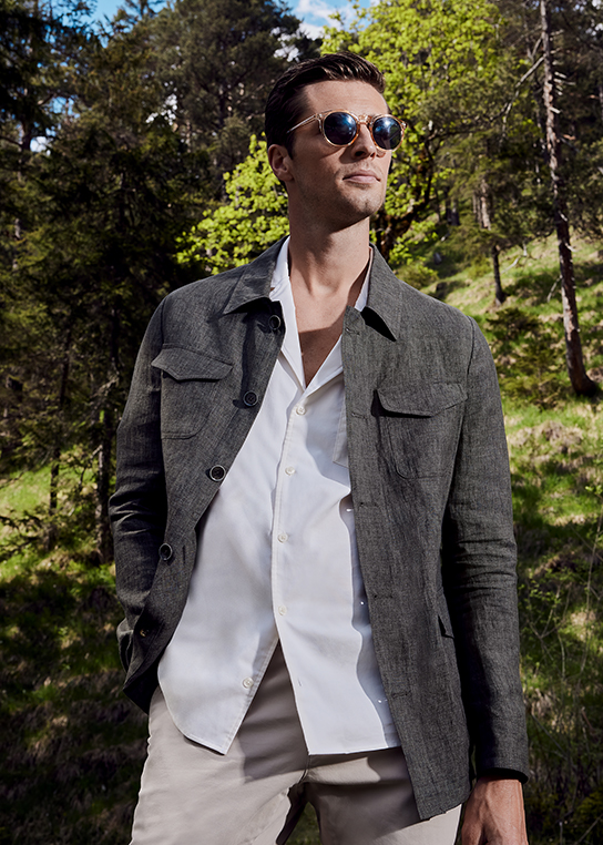 Young man with brown hair in white shirt, light beige trousers and brown open linen jacket. He is also wearing round sunglasses. A forest backdrop is seen in the background.