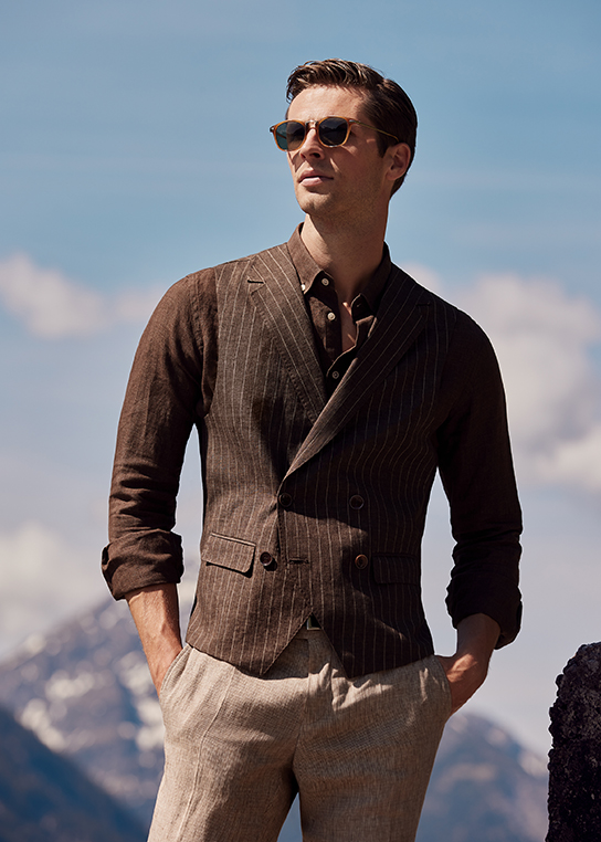 Young man with brown hair in dark brown linen shirt, brown light striped linen waistcoat and beige trousers. He also has both hands in his trouser pockets and wears sunglasses. A mountain backdrop can be seen in the background.
