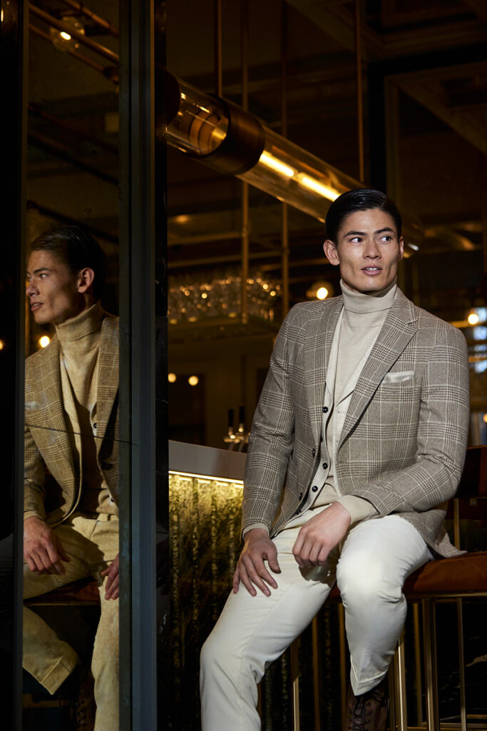 Young man with brown hair wearing a light beige turtleneck jumper and an open beige and white checked jacket. He is wearing white trousers with it. He is sitting on a bar stool at a bar.