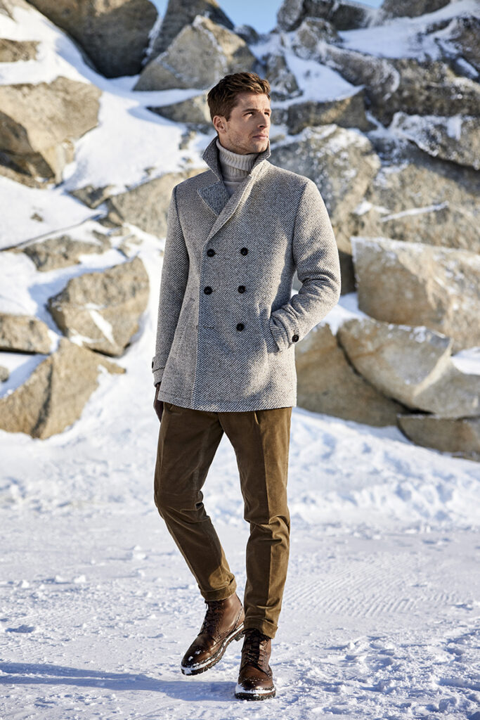 Young man with brown hair in grey double-breasted closed coat, light-coloured turtleneck jumper, brown corduroy trousers and brown shoes. In the background you can see rocks covered with snow.