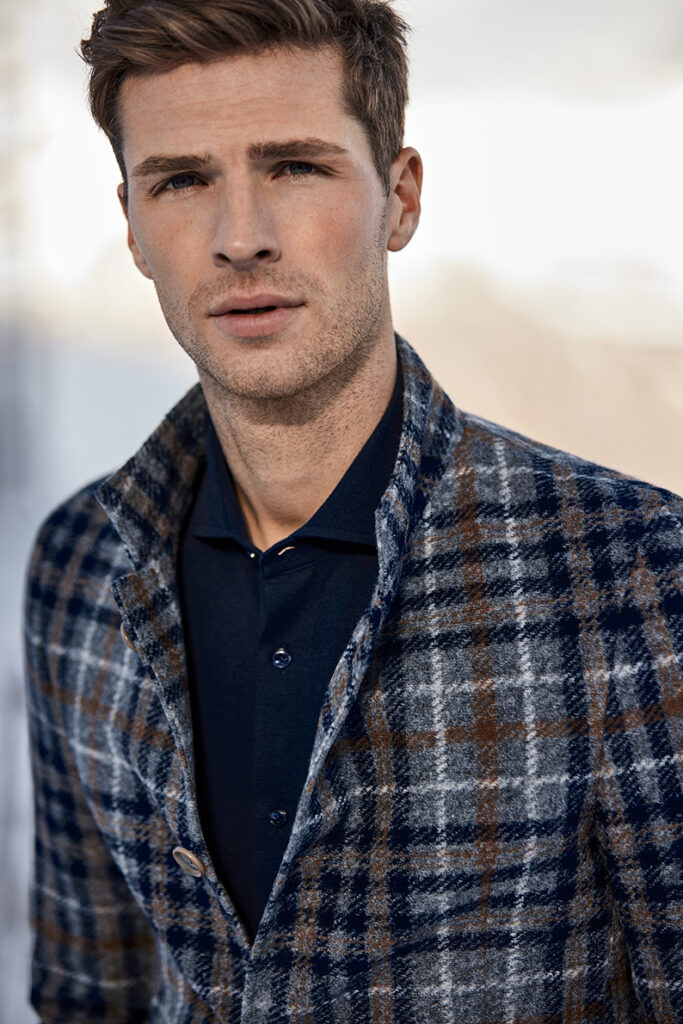 Young man with brown hair in brown-blue-white checked jacket in coarse tweed look and blue shirt underneath. This is a close-up photograph.