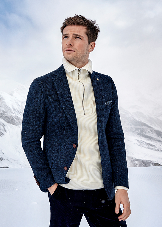 A young man with brown hair wearing a coarse white turtleneck jumper and our blue Harris Tweed limited edition herringbone jacket over it. He is wearing blue trousers with it.