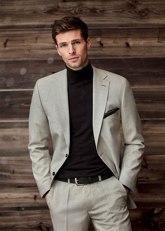 Young man with brown hair in light beige suit and black turtleneck. He stands in front of a wooden wall and looks directly into the camera.